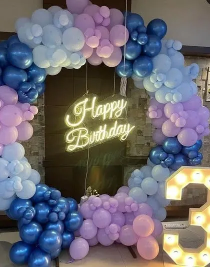 A chic and modern circular balloon frame for an intimate birthday celebration at home. This setup combines shades of blue and lavender balloons with a central 'Happy Birthday' neon sign and a large light-up number '3'.