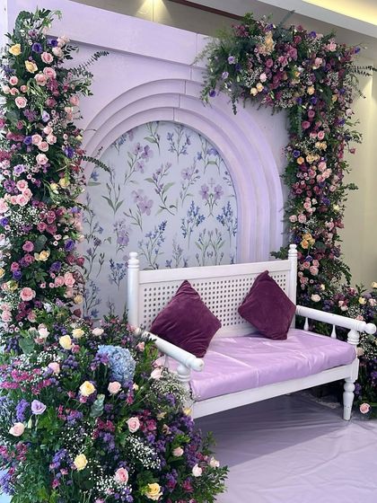 A full view of the lavender-themed Aqeeqah decor for Samadh. The setup includes a white bench for photos, set against a floral-patterned backdrop and framed with gorgeous purple flower arrangements.