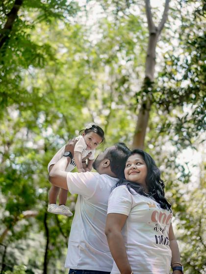 A family of three in a park, with the father lifting their child up high towards the sky.