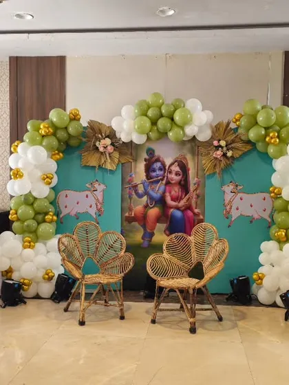A complete Krishna-themed baby shower stage I created. This playful decor features a custom backdrop with Radha and Krishna, framed by an olive green and white balloon arch, and includes themed elements like cow cutouts, wicker chairs, and 'BABY' blocks for a perfect photo opportunity.