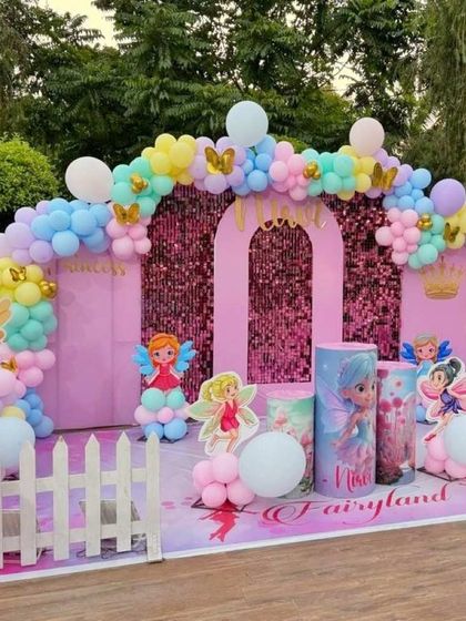A beautiful Fairyland-themed birthday setup for an outdoor party. It features a pink castle backdrop with a shimmer panel, fairy cutouts, themed pedestals, and a pastel balloon arch.
