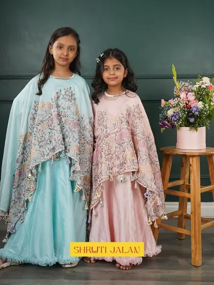 Two sisters in matching cape gowns, one in sky blue and one in blush pink. The floral printed capes with tassels are the highlight of these beautiful outfits.