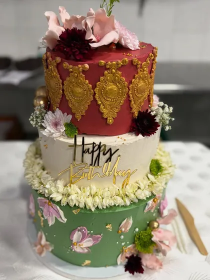 A three-tier cake with a touch of Indian royalty. The design combines a rich maroon tier with gold detailing, a classic white tier, and a base of green palette-knife florals.
