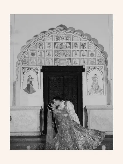 A romantic black-and-white photo of the groom kissing the bride's forehead in front of an ornate, traditional doorway.