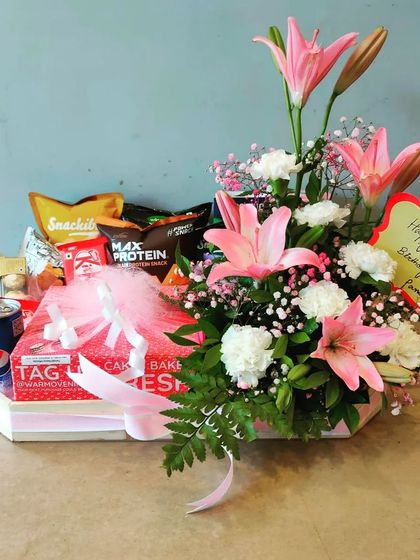 A bright and cheerful hamper featuring a beautiful arrangement of pink lilies and white carnations. It's paired with snacks, a drink, and a cake box, making it a complete celebration package.