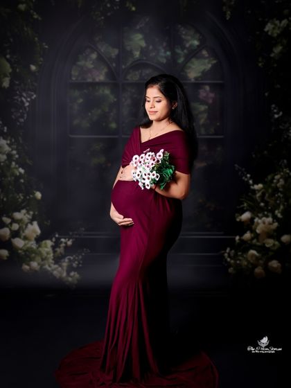 A moody and elegant solo portrait. The mom-to-be holds a simple bouquet, her dark red gown standing out against the dark, painterly floral backdrop.