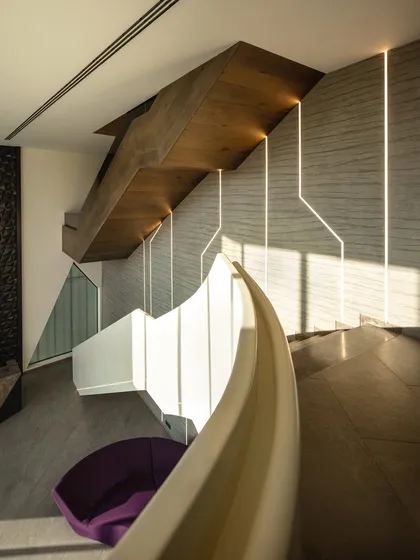 In this office building, the grand staircase features a curved white balustrade that contrasts with the textured grey walls and wood-clad underside. Integrated lighting follows the lines of the wall, creating a dynamic and welcoming entrance.