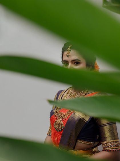 An artistic shot of a bride peeking through green leaves. Her orange and navy blue saree creates a striking contrast with the natural surroundings.