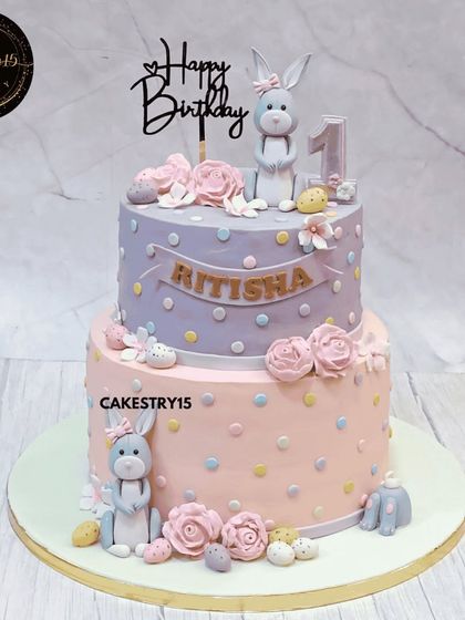 An elegant two-tier blueberry cake for a first birthday, featuring a classy bunny theme. The soft pastel colors, polka dots, and sweet fondant bunnies create a charming and sophisticated design.
