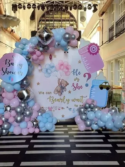 A large-scale "He or She, We can Bearly wait" baby shower theme in a spacious hall. The setup includes a round backdrop, pink and blue balloon arrangements, and themed props like "BABY" blocks.