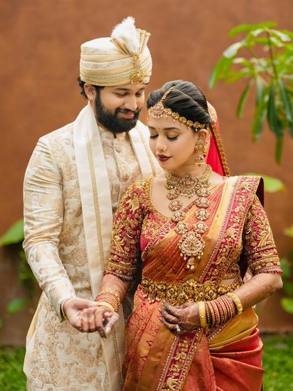 A couple stands together in their wedding finery. Her red and gold bridal saree with an intricately embroidered blouse is perfectly paired with his cream and gold sherwani.