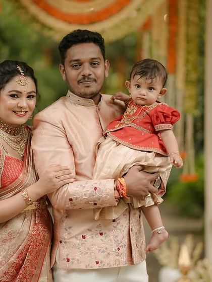 A classic family portrait. The mother's red and cream saree, the father's embroidered peach jacket, and the child's matching red and cream outfit were all designed to create a cohesive and beautiful family look.