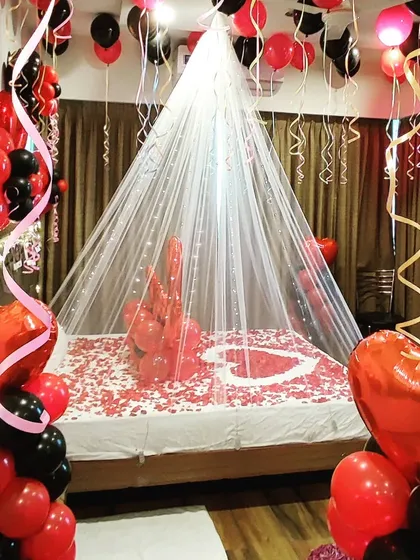 A classic romantic room setup in a red and black color scheme. The bed is decorated with rose petals and balloons under a sheer, lit canopy, creating a perfect surprise for a loved one.