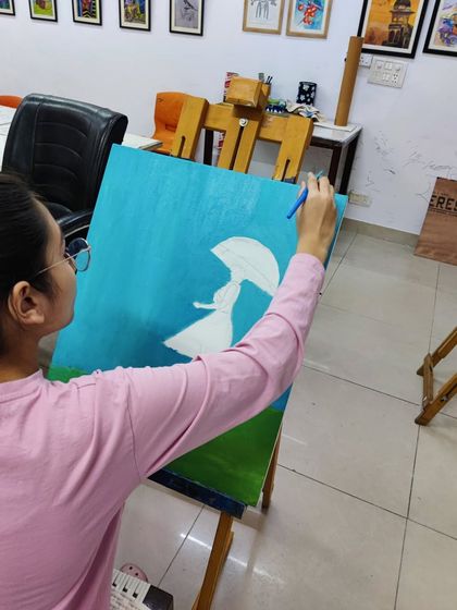 A student works on an imaginative painting of a girl with an umbrella in a field, using a palette knife and brush to create texture.