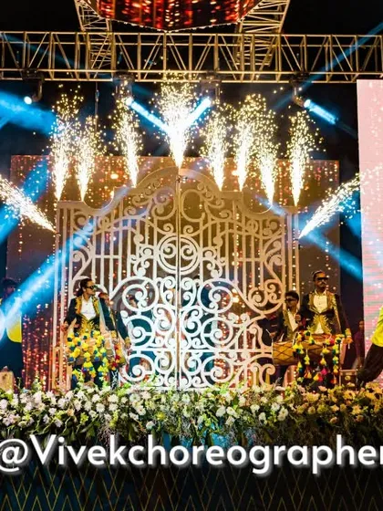 The stage is set for a grand performance. Dancers in coordinated outfits perform in front of large gates, with cold pyros adding to the drama before the couple's entry.
