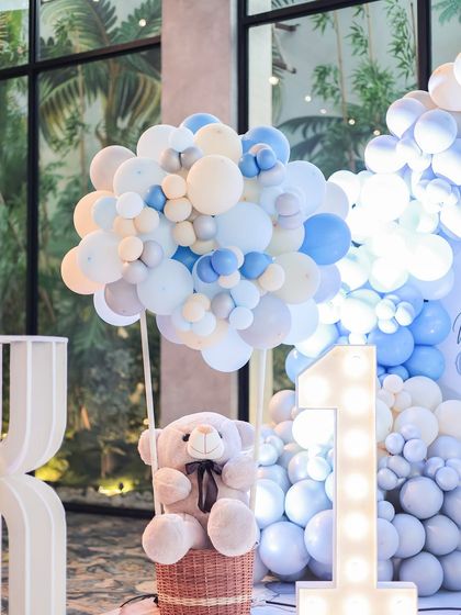 A teddy bear sits in a basket lifted by a large cluster of blue, white, and silver balloons, resembling a hot air balloon. This creative decor piece is paired with a light-up number '1' for a first birthday party.