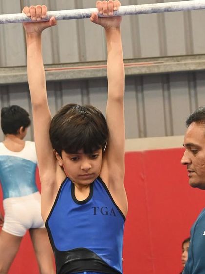 A young gymnast shows intense focus while training on the bars, with his coach watching closely.