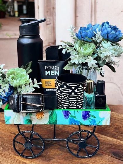This charming gift hamper for him is arranged in a decorative miniature cart. It includes men's grooming essentials, a stylish belt, perfume, and a patterned mug, accented with blue floral touches.