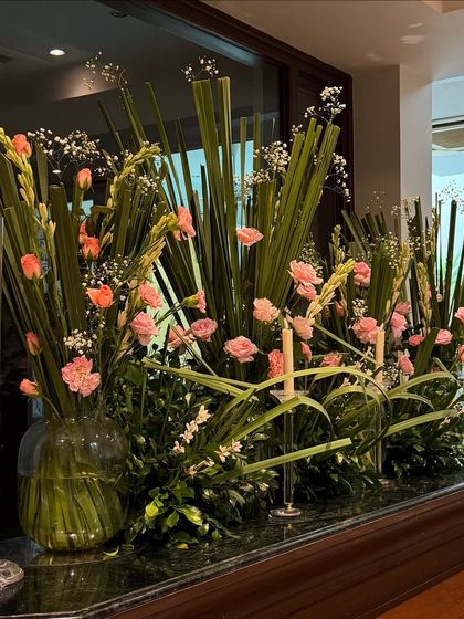An elegant floral arrangement of pink carnations and tall green foliage, part of the decor for an 80th birthday. We use fresh, high quality flowers to bring life and color to every corner of the event.