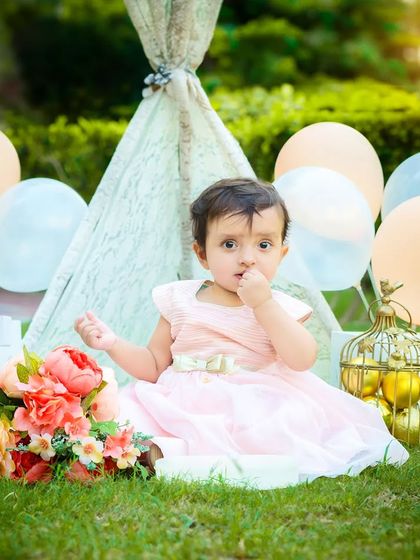 A baby girl celebrates her first birthday with an outdoor party, sitting in front of a lace teepee decorated with balloons and flowers.