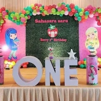 A "Berry First Birthday" party with a Strawberry Shortcake theme. The setup includes a grass wall, character cutouts, and large 'ONE' letters.