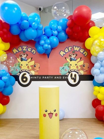 A Pokemon themed birthday decoration for two brothers. The shared backdrop features Ash, Pikachu, and other characters, framed by a colorful balloon arch.