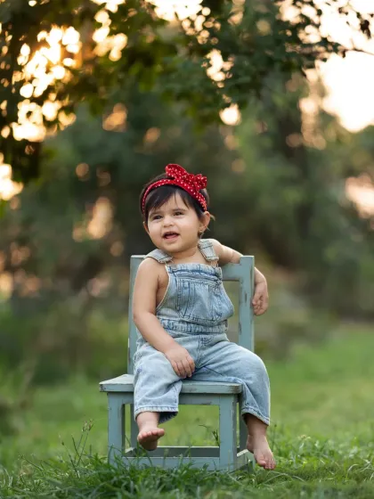 A little girl in overalls and a red bandana sits on a chair in a green field. A classic and cute look for an outdoor kids photoshoot.