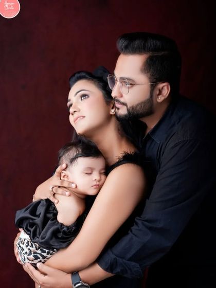 A classic family portrait against a deep red backdrop. The parents hold their sleeping baby close, creating an image of warmth, elegance, and love.