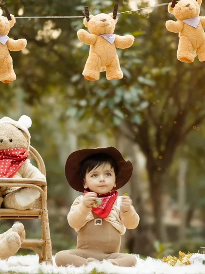 A sweet portrait of a baby in a cowboy hat, sitting among teddy bears in a green, natural environment. The soft focus on the background makes the adorable subject stand out.