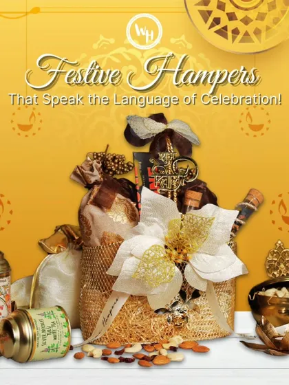 A festive hamper that speaks the language of celebration. This gift is arranged in a golden jute bag and includes tins of fine tea, nuts, and other treats, adorned with a large white and gold flower.
