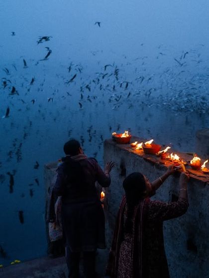 Devotees light diyas on the banks of the Yamuna river in Delhi during the blue hour. A flock of birds fills the sky, creating a magical and serene atmosphere.