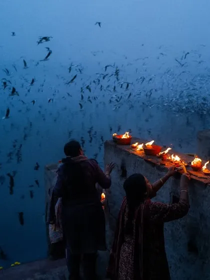 Devotees light diyas on the banks of the Yamuna river in Delhi during the blue hour. A flock of birds fills the sky, creating a magical and serene atmosphere.