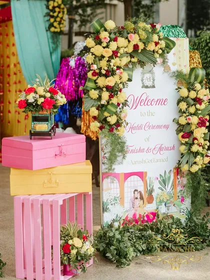 A personalized welcome sign for the Haldi ceremony, featuring a custom caricature of the couple. The sign is framed with a lush arch of yellow and red flowers.