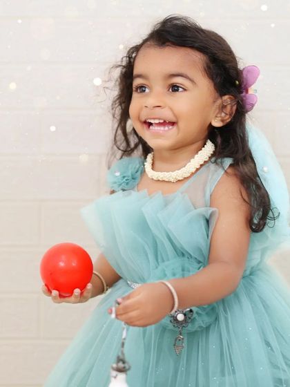 A smiling girl in a light blue dress holds a red ball, her joy shining in the natural light. Outdoor portraits like this are full of life and genuine expression.