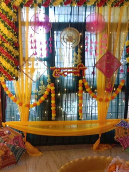 A creative Haldi setup for a home window area. The decor uses yellow drapes, marigold garlands, and colorful hangings to create a festive frame.