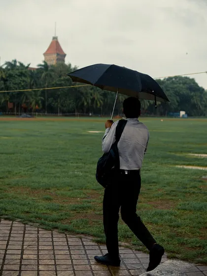 A man in formal wear walks along a paved path in a park, holding an umbrella. It's a slice of the daily commute for many Mumbaikars during the rainy season.