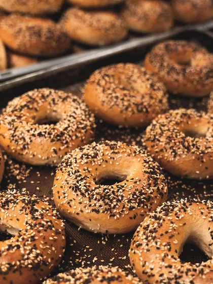 A fresh batch of our hand-rolled bagels, straight from the oven. We boil them before baking to achieve that classic shiny crust and dense, chewy interior. Topped generously with sesame seeds, they are a breakfast favorite.