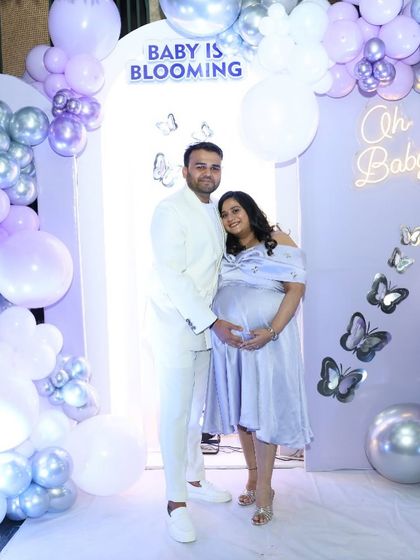The happy couple posing in front of their 'Baby is Blooming' backdrop. The lavender and silver color scheme is both soft and celebratory.