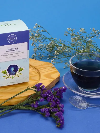 A styled product shot for Darjeeling Sapphire Tea, featuring the packaging alongside a cup of the distinctive blue tea. The composition is enhanced with complementary blue and white floral props to create an elegant theme.