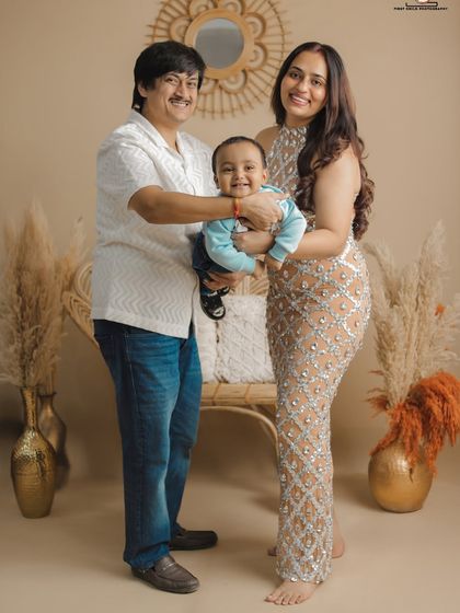 A full-length family portrait showcasing their coordinated style. The parents proudly present their smiling son in a chic, boho-inspired studio setting with pampas grass and rattan furniture.