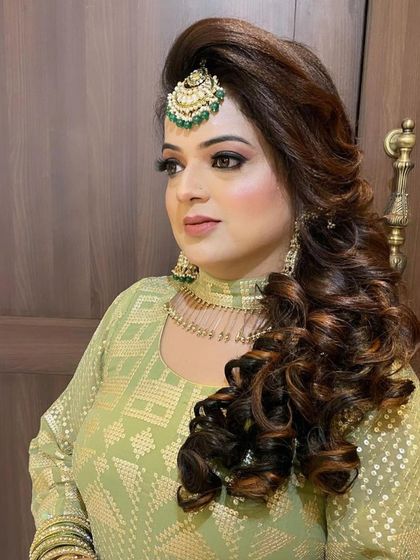 A regal bridal look featuring a voluminous, curled side-hairstyle and a statement maang tikka. The makeup is bold and beautiful, perfect for a bride.