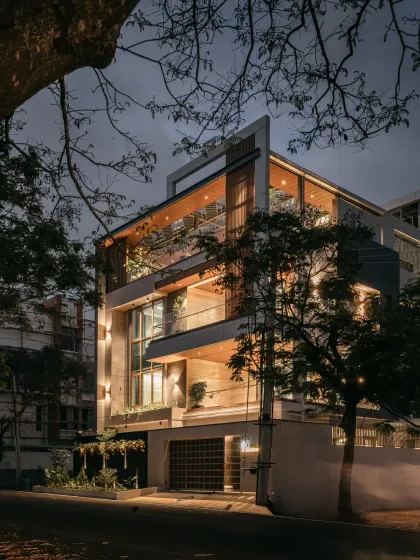 A corner view of the Hennur residence at night, framed by trees. This angle shows how the different volumes of the house come together, with the lighting scheme emphasizing the modern architectural form.
