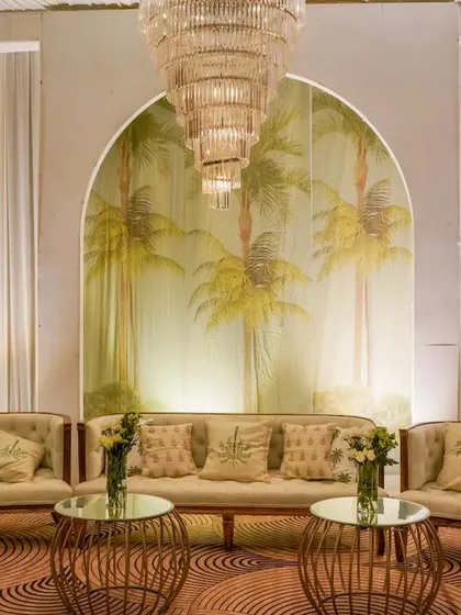 A tropical-themed lounge area for a reception, featuring a hand-painted palm tree mural, comfortable beige seating with themed cushions, and elegant glass-topped tables.