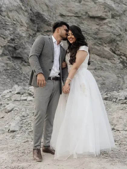 A sweet and affectionate moment against a rugged, natural backdrop. He kisses her on the cheek as she smiles, holding his hand. This full-length shot captures their joy and the beautiful contrast of her white dress against the rocks.