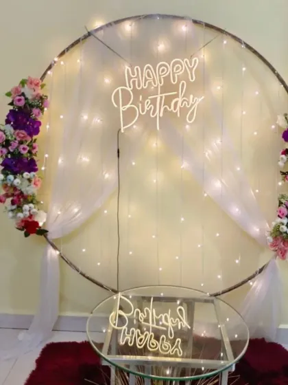 A circular frame with fairy lights and white drapes serves as a simple and elegant backdrop. The floral accents and "Happy Birthday" neon sign complete the look.