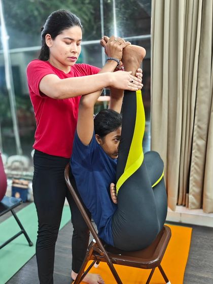 An instructor provides careful assistance in a seated leg extension using a chair. This method helps improve hamstring flexibility while maintaining proper spinal alignment.