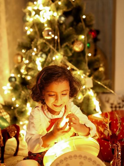 The magic of Christmas lights. This little girl's face is lit up with joy as she plays with the warm, glowing lights in front of the tree.