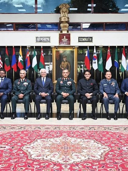 A formal group photograph with senior officers and fellow speakers at the Defence Services Staff College. The flags of various nations in the background represent the international nature of the audience.