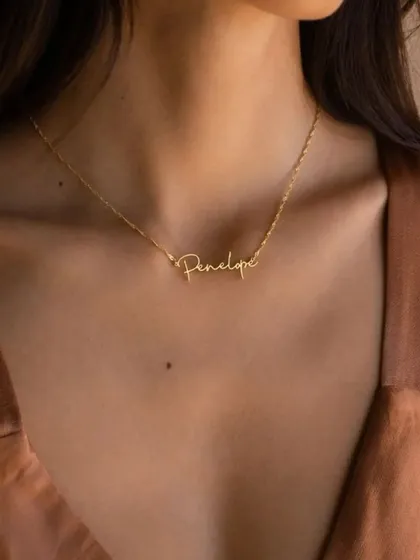 The 'Penelope' script necklace worn against a simple top, showing how it can be an elegant centerpiece for any outfit. The fine chain and polished script create a look of effortless sophistication.