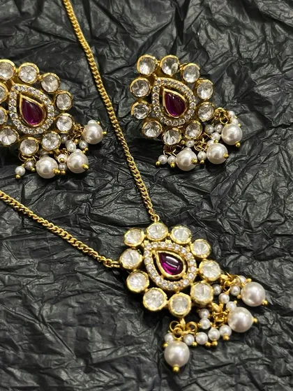 A stunning gold-plated necklace set. This piece features a pendant and matching earrings adorned with a central ruby-red stone, surrounded by white Kundan and delicate pearl drops.